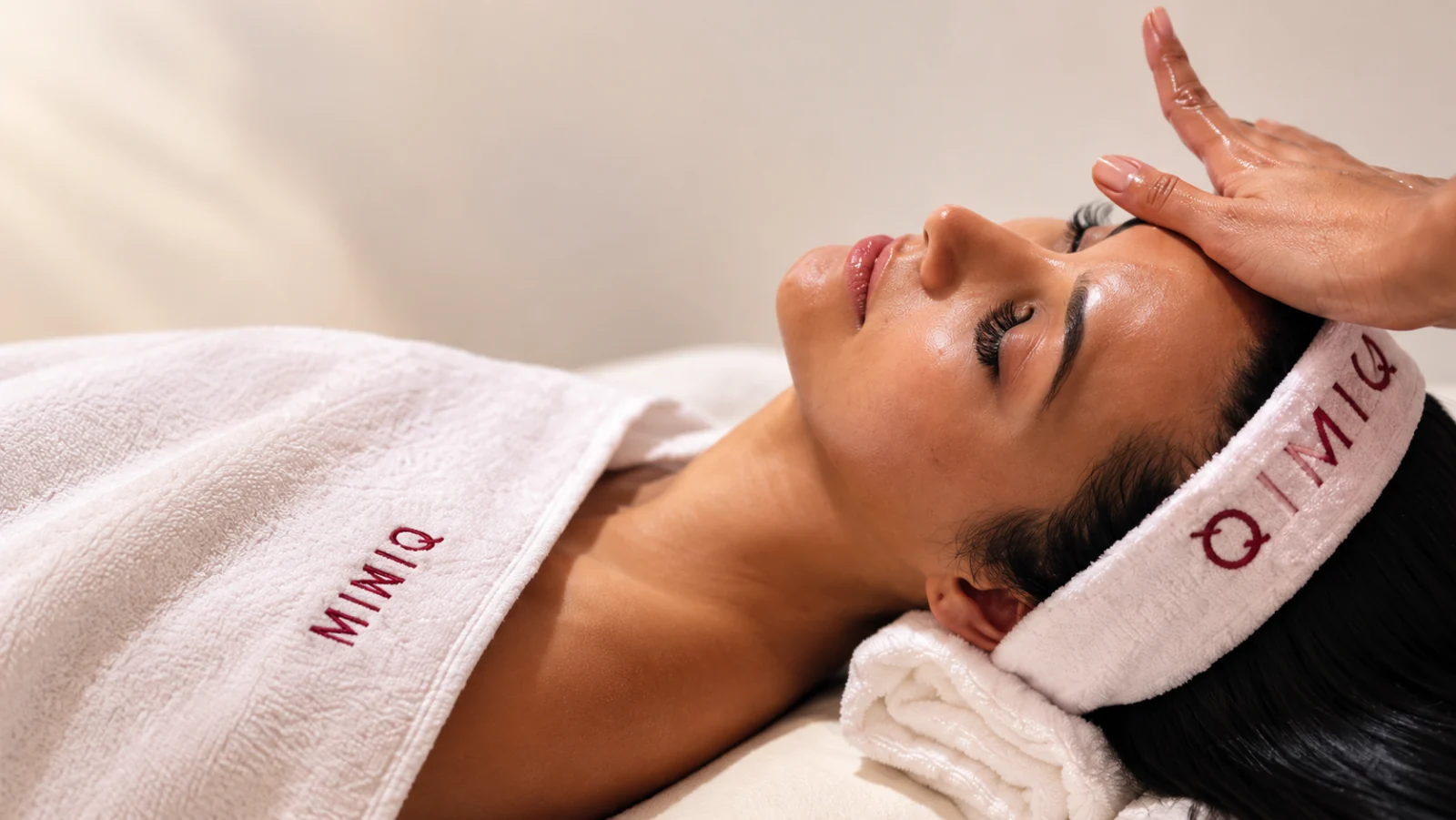 MIMIQ facial massage with a signature white headband and towel, in a source-based editorial image.